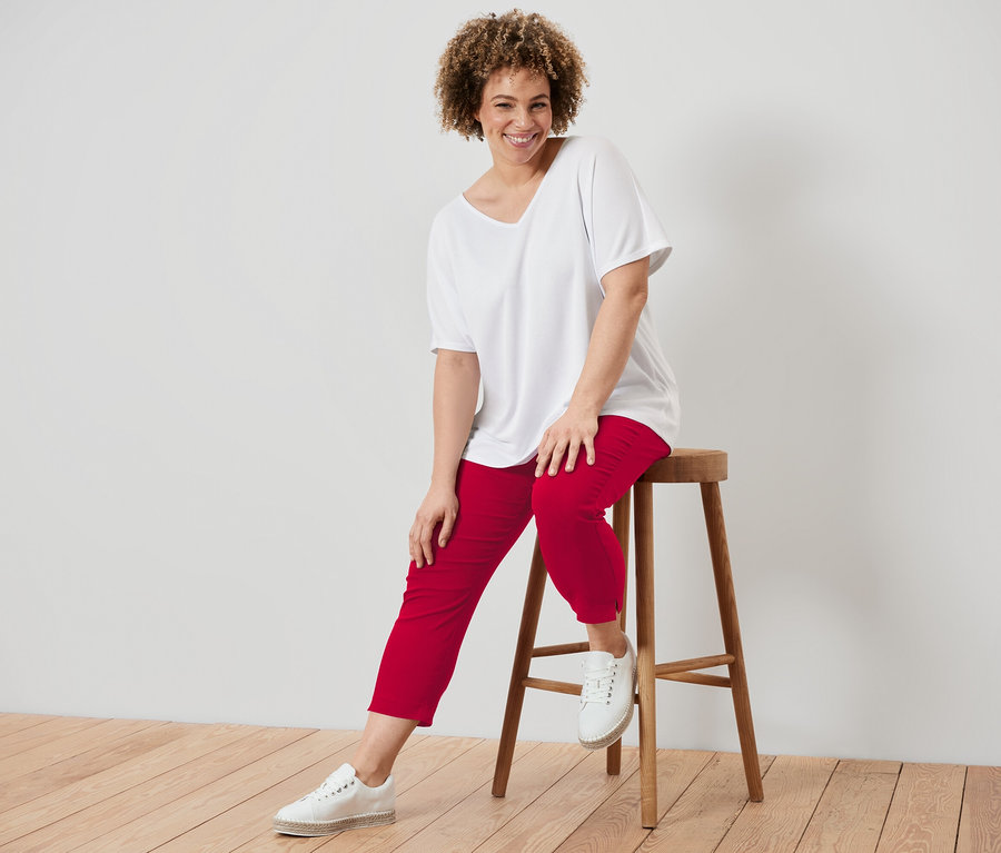 Frau mit lockigem Haar sitzt auf einem Holzschemel, trägt ein weißes T-Shirt und rote Hosen.