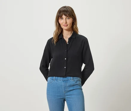 Frau trägt eine schwarze Langarmbluse und blaue Jeans vor einem weißen Hintergrund.