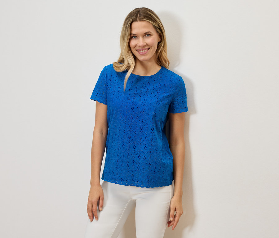 Eine Frau lächelt und trägt ein blaues, kurzärmeliges Lochmuster-T-Shirt und eine weiße Hose vor einem weißen Hintergrund.