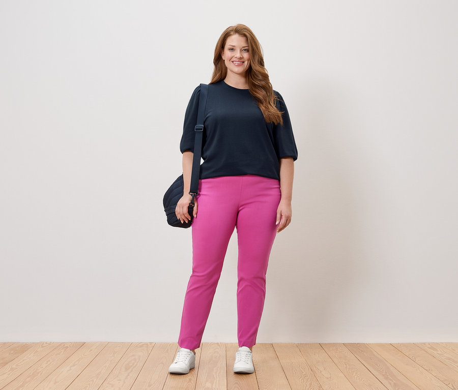 Frau steht in marineblauem Oberteil und pinkfarbener Hose mit einer Umhängetasche vor einer weißen Wand.