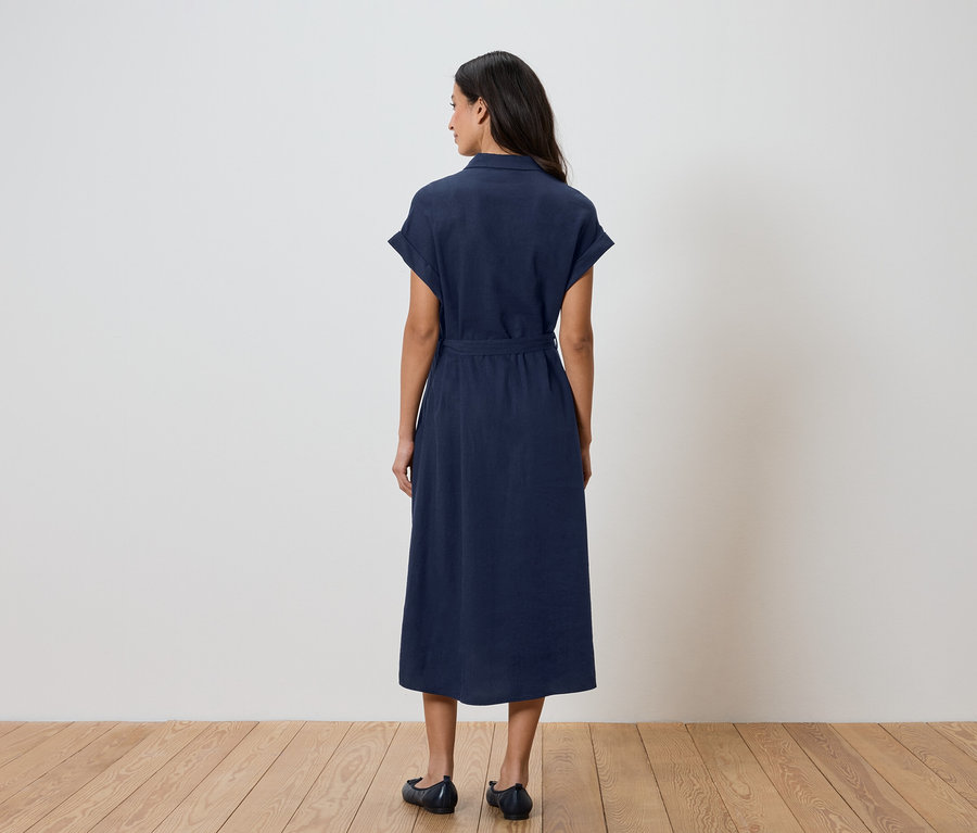 Eine Frau von hinten in einem marineblauen Hemdkleid mit Gürtel und flachen Schuhen vor einem weißen Hintergrund.
