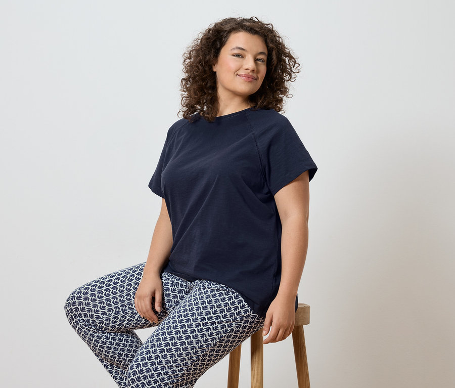 Frau mit lockigem Haar sitzt auf einem Hocker und trägt ein marineblaues T-Shirt und gemusterte Leggings.