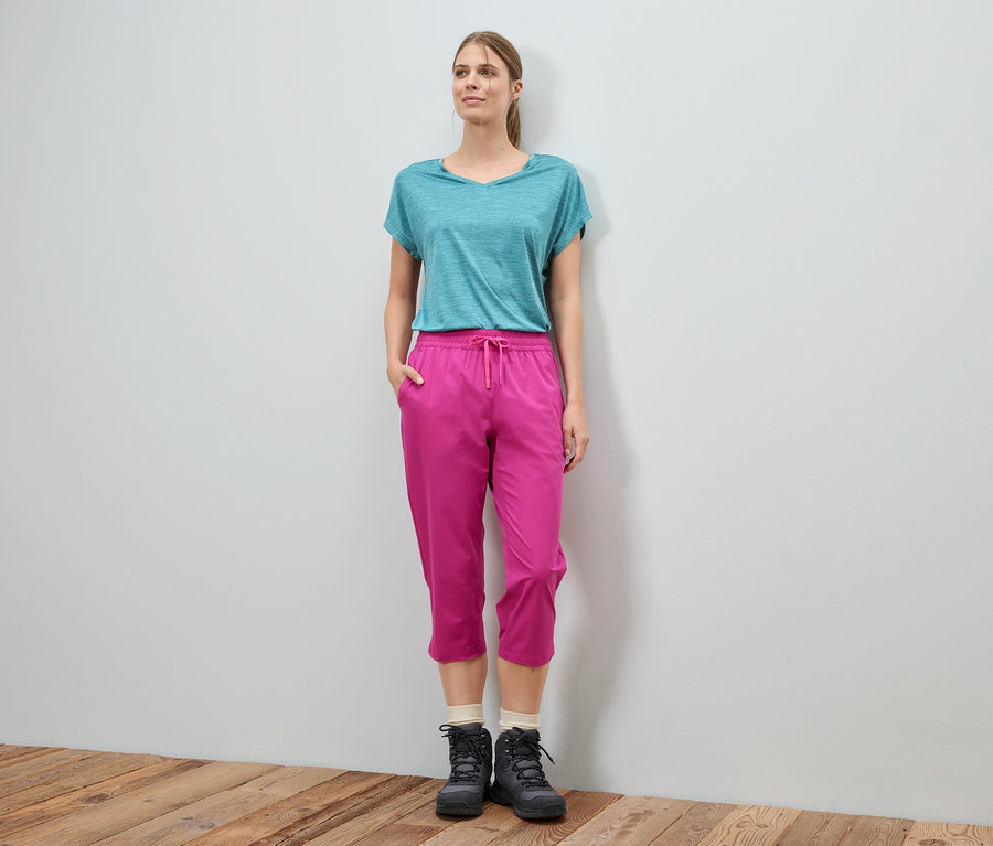 Frau steht vor einer Wand und trägt ein blaues T-Shirt, pinke Hose und schwarze Stiefel.