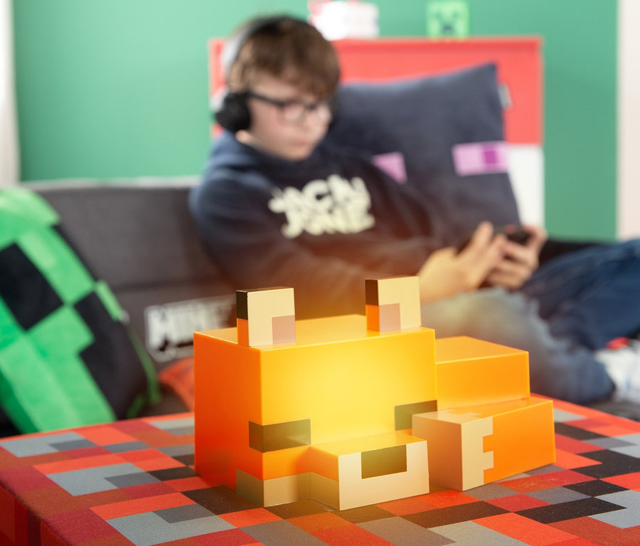 Ein Jugendlicher mit Kopfhörern sitzt auf einem Sofa und schaut auf sein Handy. Im Vordergrund steht eine Minecraft-Fuchs-Lampe auf einem Tisch.