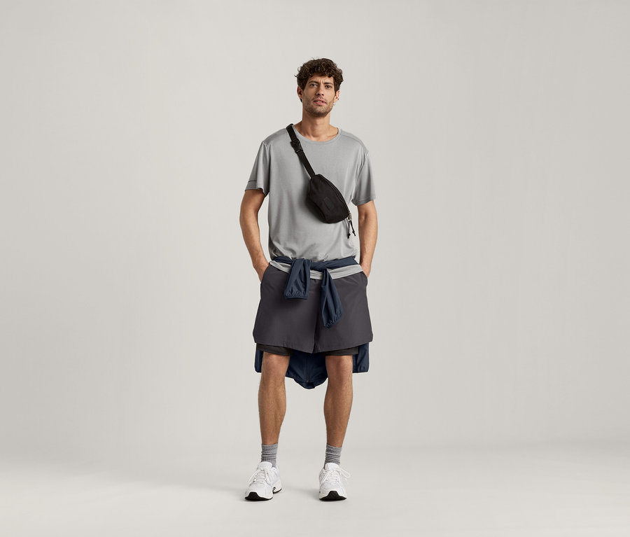 Mann trägt ein hellgrau meliertes Sportshirt, eine schwarze Bauchtasche, Shorts und eine Jacke um die Hüfte.