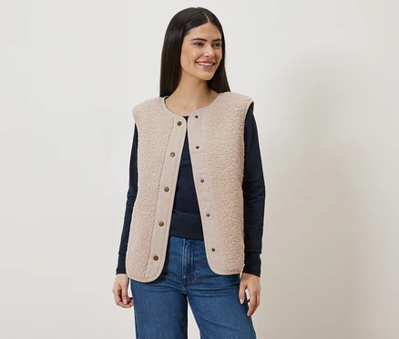 Frau mit langem, dunklem Haar trägt eine ärmellose, hellbraune Teddyfellweste über einem marineblauen Langarmshirt und Jeans.