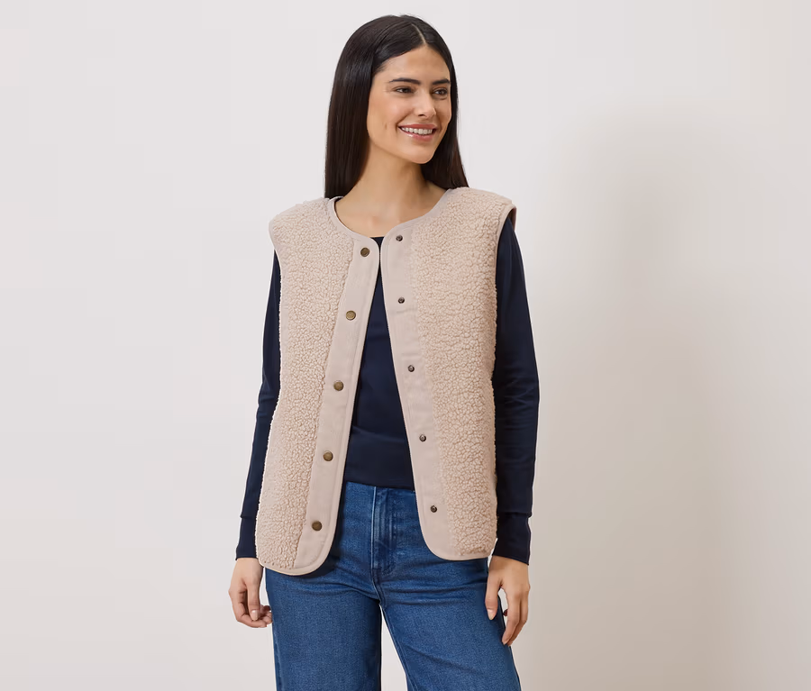 Frau mit langem, dunklem Haar trägt eine ärmellose, hellbraune Teddyfellweste über einem marineblauen Langarmshirt und Jeans.