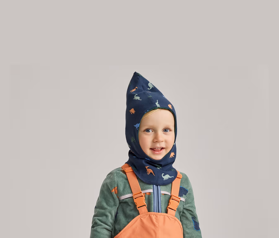 Lächelnder Junge mit blauem Dino-Balaclava, grünem Fleecepullover und orangefarbener Latzhose vor grauem Hintergrund.