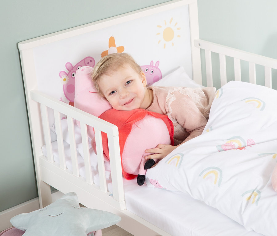 Ein kleines Mädchen liegt in einem weißen Kinderbett und kuschelt mit einem Peppa Pig-Kissen.