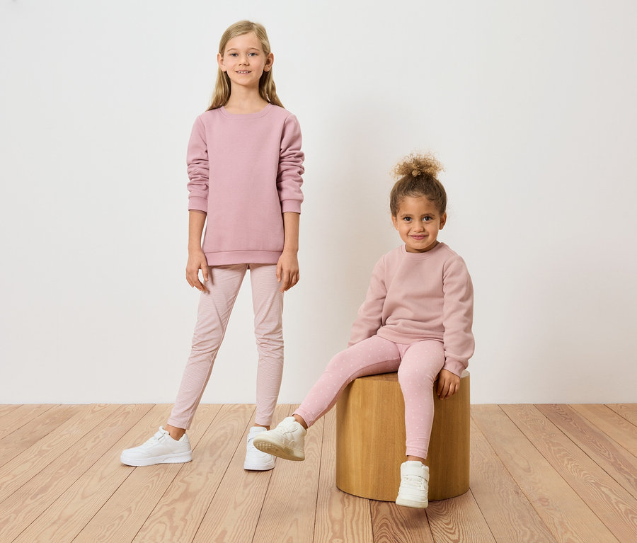 Zwei Mädchen, eines stehend und eines auf einem Hocker sitzend, tragen passende rosa Sweatshirts und Leggings.