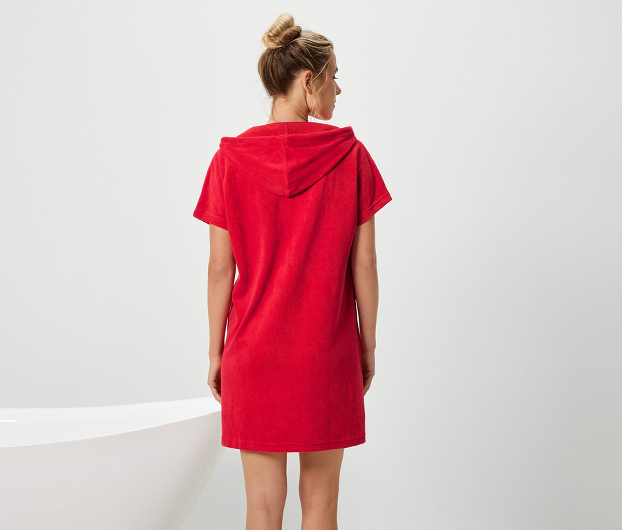 Eine Frau mit Dutt trägt ein rotes, kurzärmeliges Kapuzenkleid, mit Blick auf eine Badewanne.