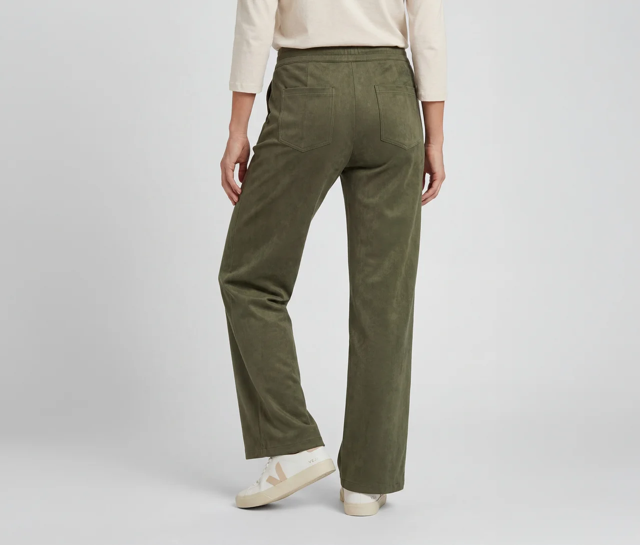 Eine Person von hinten trägt eine olivgrüne Hose mit weicher Textur und Gesäßtaschen, ein helles Langarmshirt und weiße Sneaker mit beigen Akzenten.