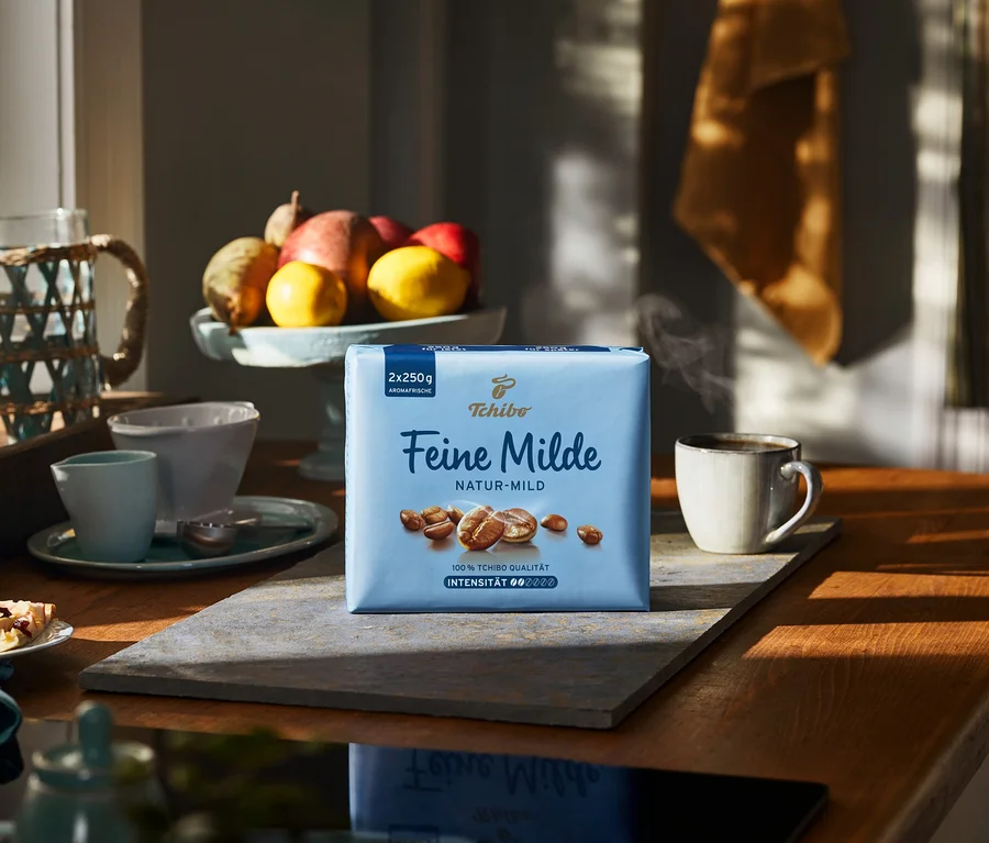 Eine Packung Feine Milde - 500 g Gemahlen steht auf einem Holztisch neben einer Tasse heißem Kaffee.