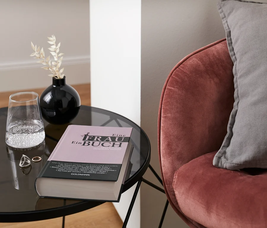 Buch „Eine Frau – ein Buch“ liegt auf einem Tisch neben einem Wasserglas und einer Vase. Im Hintergrund ist ein Sessel mit Kissen zu sehen.