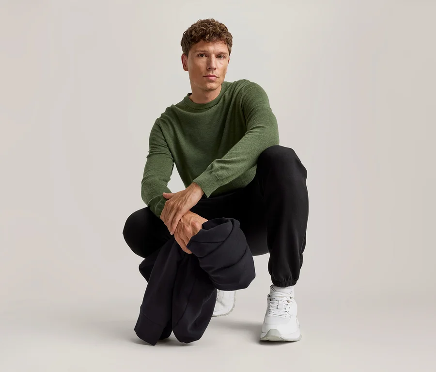Mann hockt, trägt einen grünen Langarmpullover, schwarze Jogginghose und weiße Sneaker.