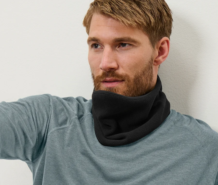 Ein Mann sitzt angelehnt an einer Wand und trägt einen schwarzen Nackenwärmer, blaue Jeans und ein blau-graues Langarmshirt.