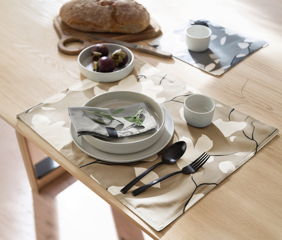 Ein heller Holztisch ist mit zwei Tischsets gedeckt. Im Vordergrund liegt ein sandfarben-florales Tischset der SCHÖNER WOHNEN-Kollektion mit einer grauen Schale, einem Teller, einer gefalteten Serviette mit grünem Zweig, schwarzem Besteck und einer kleinen Tasse. Im Hintergrund sind ein Brotlaib, ein Messer, eine Schale Mangostanen und ein graues florales Tischset mit einer Tasse zu sehen.