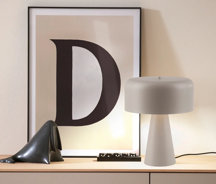 Eine beleuchtete SCHÖNER WOHNEN-Kollektion Tischleuchte »Pento« in Sandbrise steht auf einem Holztisch, daneben ein gerahmtes Poster mit dem Buchstaben D und eine Walfigur.