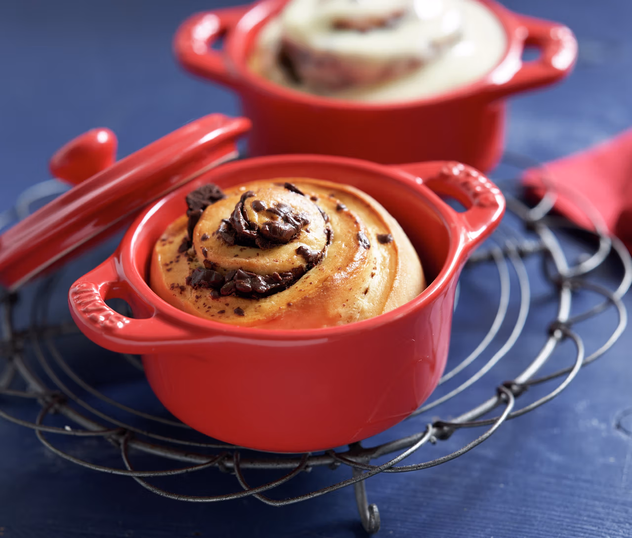 Eine kirschrote STAUB Keramik-Cocotte mit Schoko-Zimtschnecke auf einem Drahtgestell, daneben ein Deckel. Im Hintergrund eine weitere Cocotte.