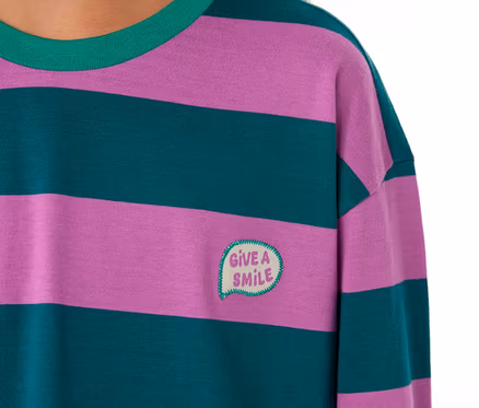 Nahaufnahme eines Kindes, das ein SCHIESSER Kinder-Pyjama Oberteil mit lila und dunkelgrünen Querstreifen, grünem Kragen und einem aufgestickten „GIVE A SMILE“-Sprechblasen-Patch trägt.