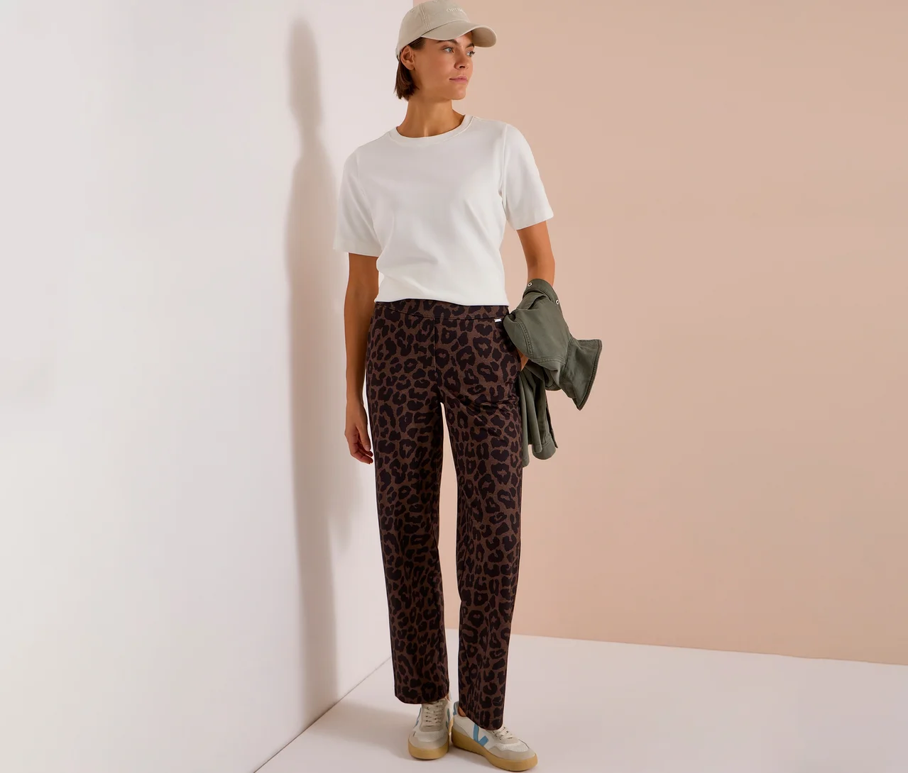 Frau trägt ein weißes T-Shirt, eine beigefarbene Kappe und eine CECIL Stoffhose mit Alloverprint, hält eine olivgrüne Jacke.