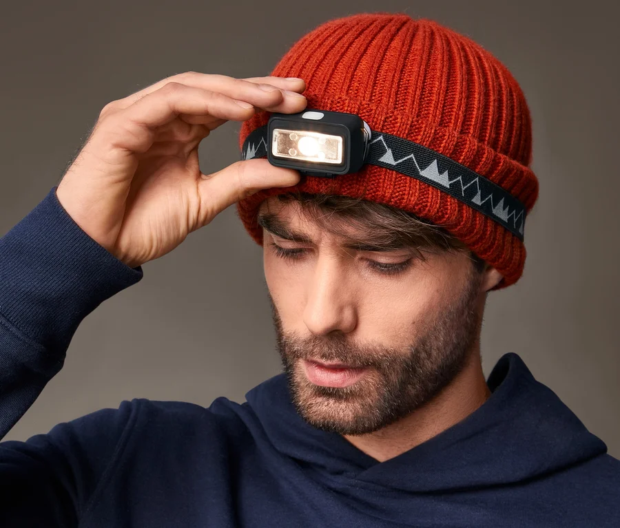 Mann justiert LED-Stirnlampe auf orangefarbener Strickmütze.