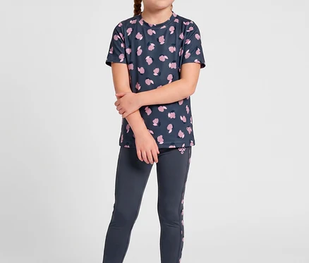 Ein junges Mädchen mit blonden Zöpfen trägt ein dunkelgraues T-Shirt mit pinken Mustern und hummel® »HMLJR AOP Gym Tights« zu pinken Turnschuhen.