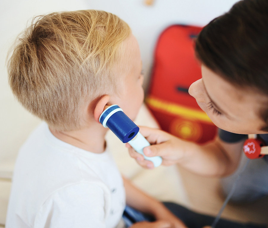 Eine Betreuerin misst die Temperatur eines kleinen Jungen mit einem Spielzeugthermometer. Im Hintergrund befindet sich ein kleiner small foot Notarzt-Rucksack.