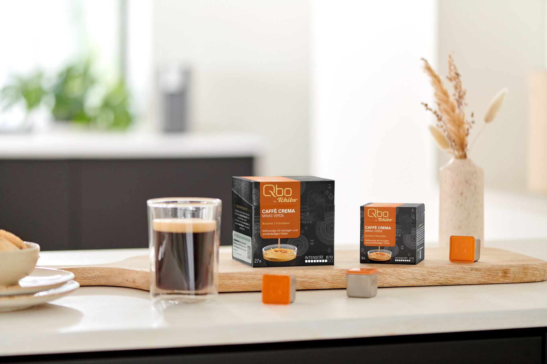 Zwei Qbo Caffè MINAS VERDE Kapselpackungen, ein Glas Kaffee und lose Kapseln auf einem Holzbrett auf der Küchenzeile, daneben Kekse und Trockenblumen.