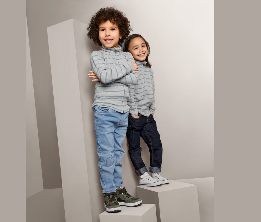 Zwei Kinder stehen auf grauen Blöcken. Der Junge links trägt eine sportive Strickjacke, Jeans und grüne Schuhe. Das Mädchen rechts trägt eine sportive Strickjacke, dunkle Jeans und weiße Schuhe.