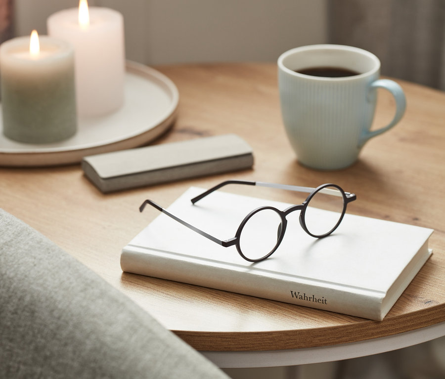 Eine ultraflache Lesebrille liegt auf einem Buch. Auf dem Tisch stehen eine Tasse und Kerzen.