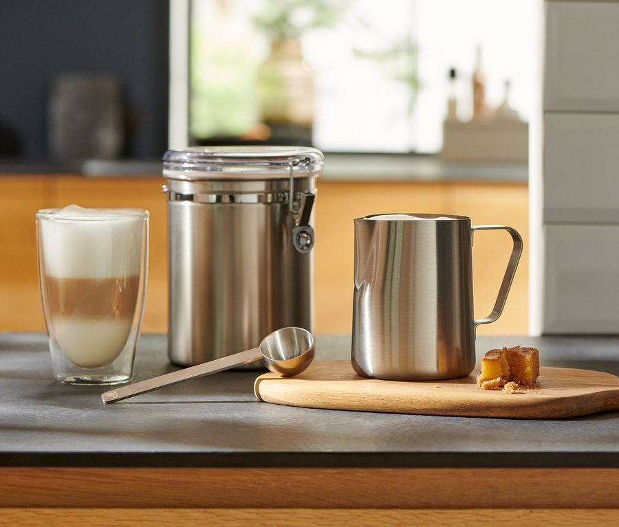 Auf der Küchentheke stehen ein Glas mit Latte Macchiato, ein silbernes Kaffeelot, ein Behälter und ein Milchkännchen.
