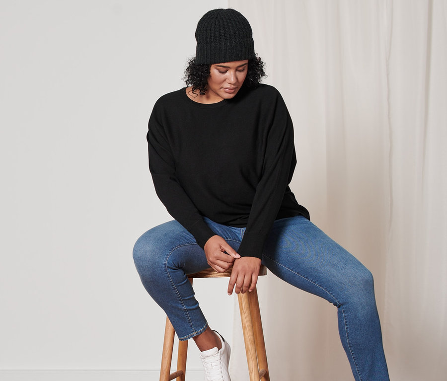 Frau sitzt auf einem Hocker und trägt einen schwarzen Feinstrick-Pullover, eine anthrazitfarbene Strickmütze mit Wolle und eine blaue Slim-Denim »Fit Emma«.