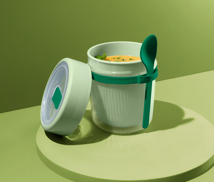 Suppenbehälter mit Suppe und grünem Löffel auf grünem Hintergrund.