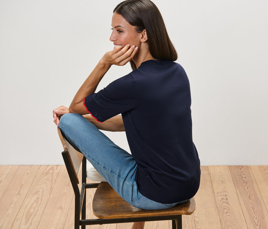 Frau sitzt auf Stuhl und trägt dunkelblauen Feinstrickpullover und blaue Jeans.