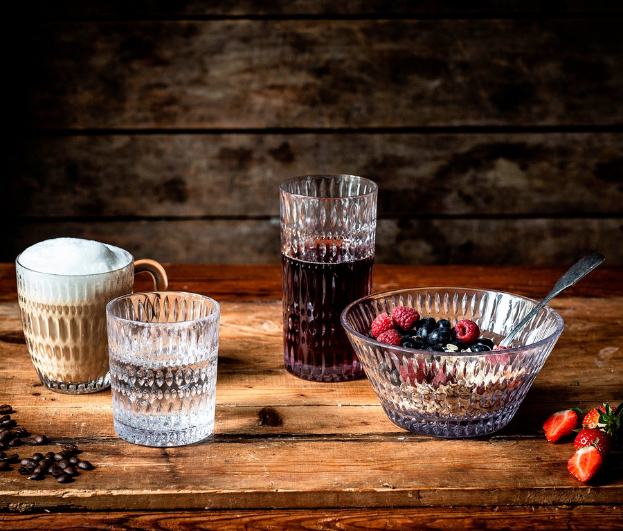Auf einem Holztisch stehen ein Glas mit Kaffee, ein Wasserglas, ein Longdrinkglas und eine Schale mit Müsli und Früchten der Marke Nachtmann Ethno.