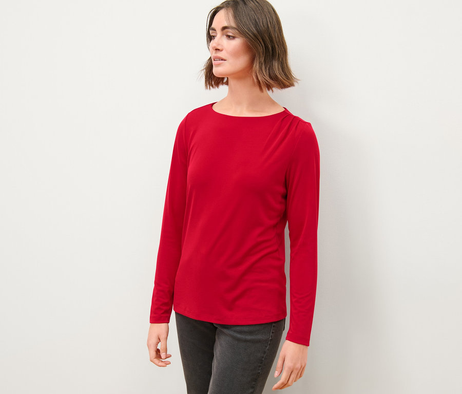 Frau in rotem Blusenshirt mit Raffung und grauen Jeans steht vor einer weißen Wand.