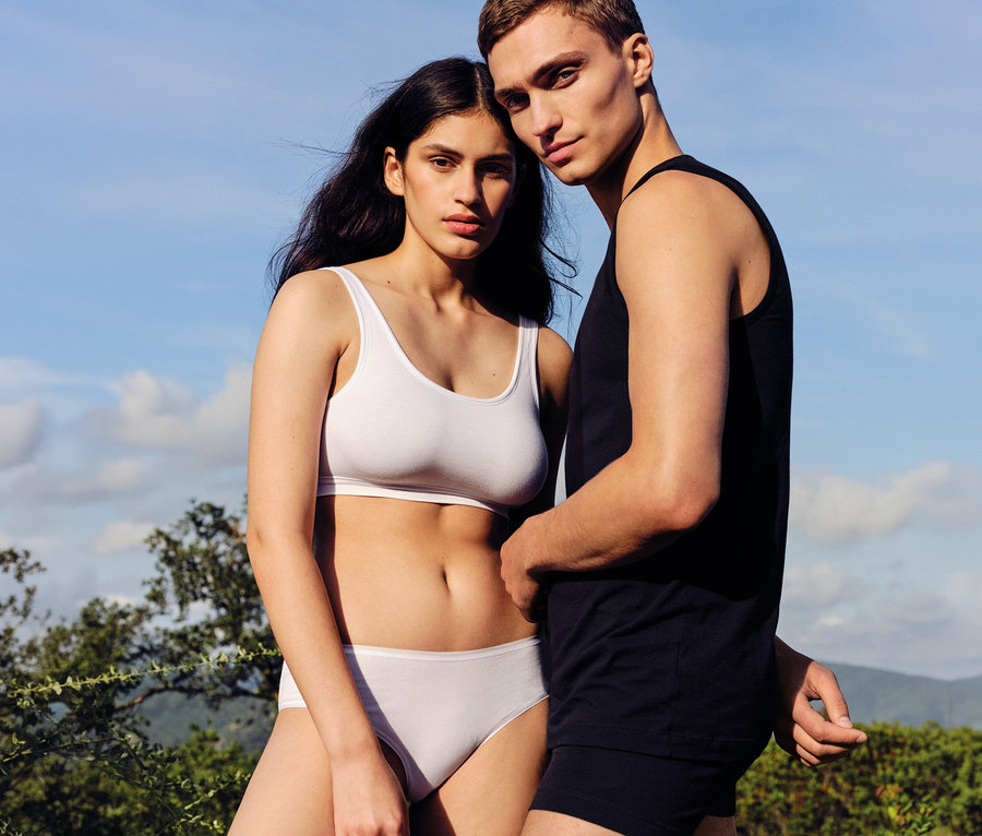 Mann und Frau stehen im Freien und blicken in die Kamera. Die Frau trägt einen weißen Sport-BH und weiße SCHIESSER Hipster. Der Mann trägt ein schwarzes Tanktop und schwarze Boxershorts.