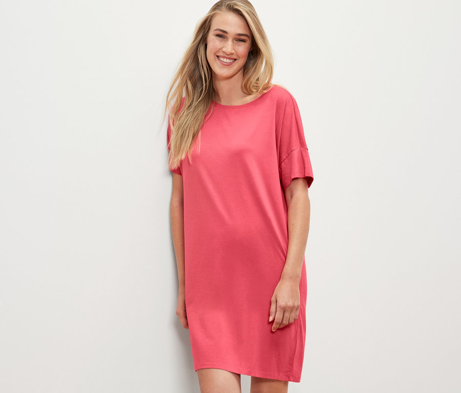 Blonde Frau im korallfarbenen Nachthemd lehnt an einer weißen Wand.