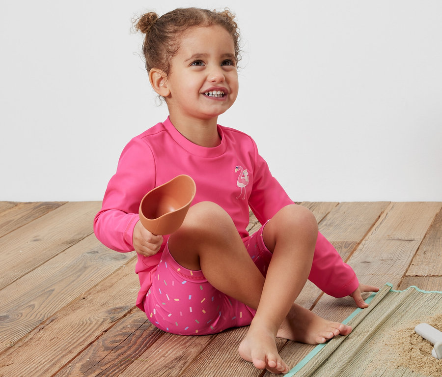Mädchen sitzt auf einer Decke und hält eine Schaufel, trägt einen pinken Kinder-Kombi mit UV-Schutzfaktor 80.