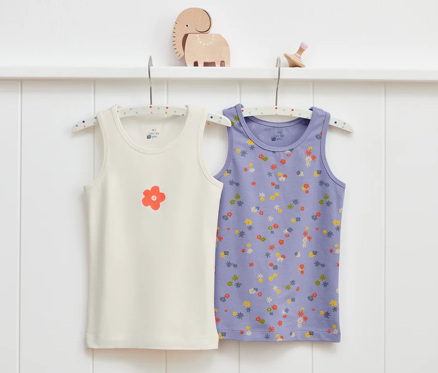 Zwei Kleinkind-Unterhemden hängen an einem Kleiderbügel an einer weißen Wand. Ein Unterhemd ist cremefarben mit einer orangefarbenen Blume, das andere ist lila mit einem Blumenmuster.