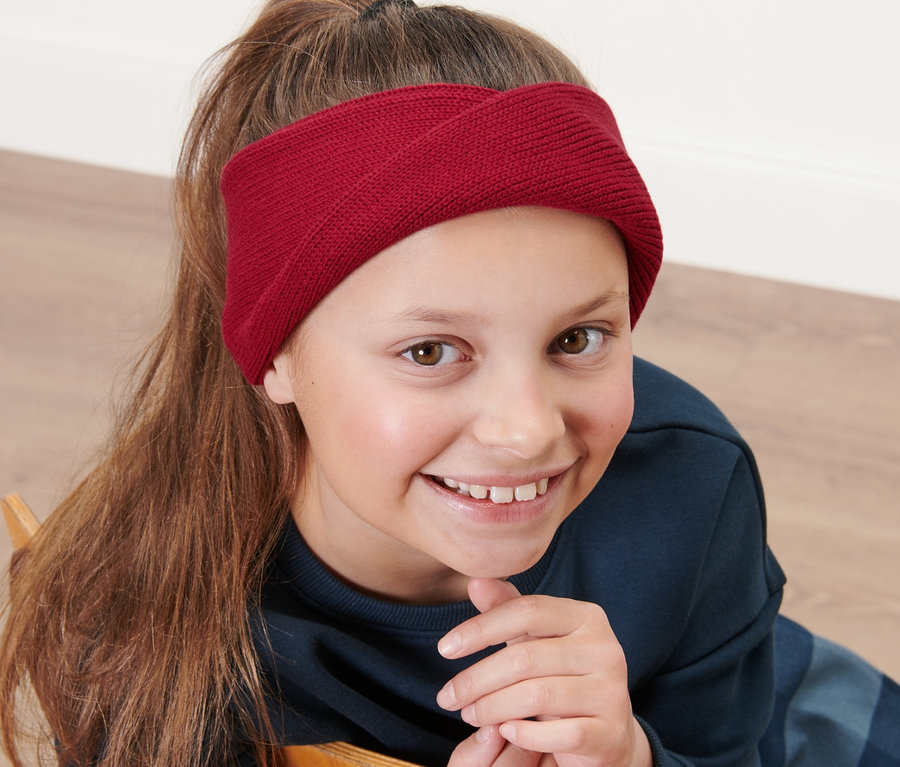 Mädchen sitzt auf einem Stuhl und trägt ein Kinder-Strickstirnband.