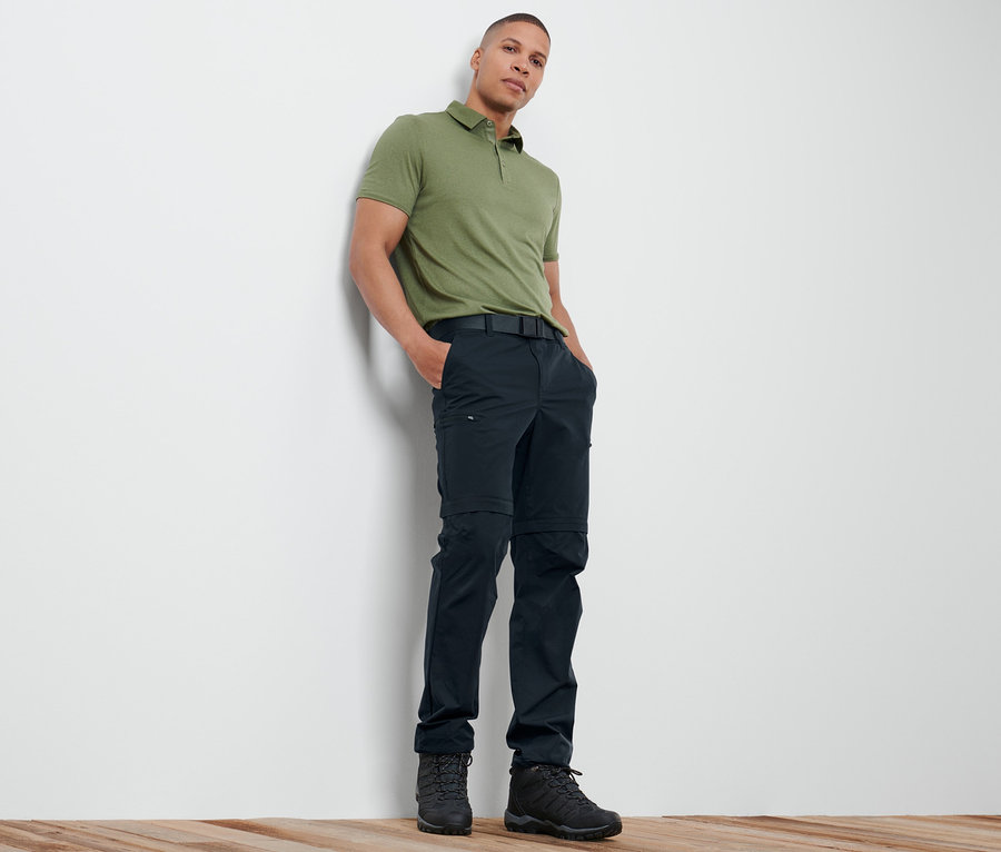 Mann lehnt an Wand, trägt grünes Poloshirt und marineblaue 2-in-1-Funktionshose.