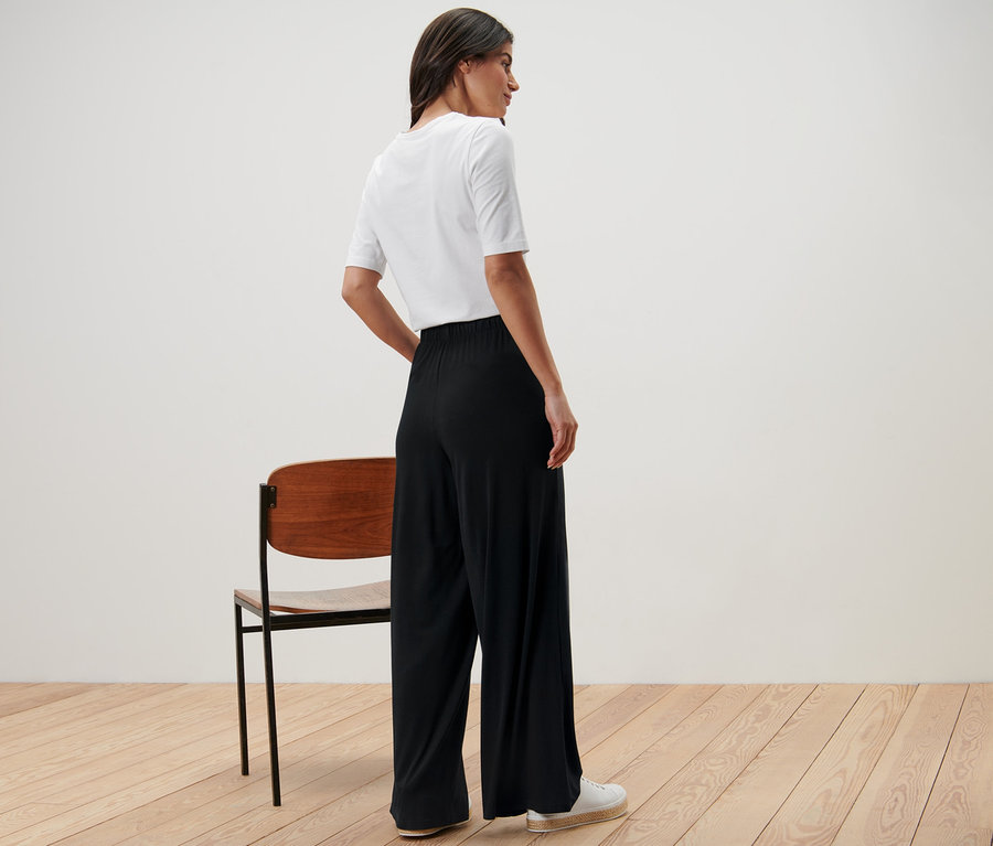 Eine Frau steht seitlich und trägt eine schwarze Palazzohose und ein weißes T-Shirt.