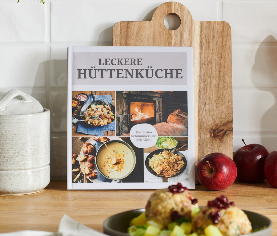 Das Buch »Leckere Hüttenküche« steht auf einem Holztisch neben drei roten Äpfeln.