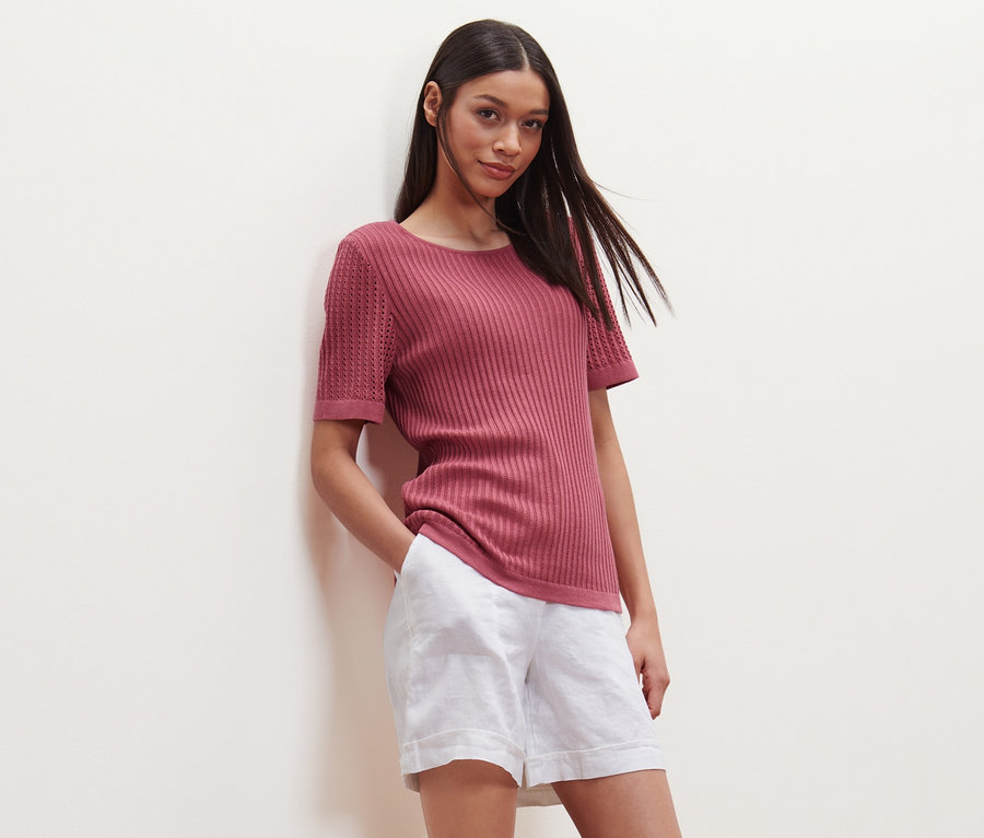 Frau posiert in beerefarbenem Strick-Shirt und weißen Shorts.