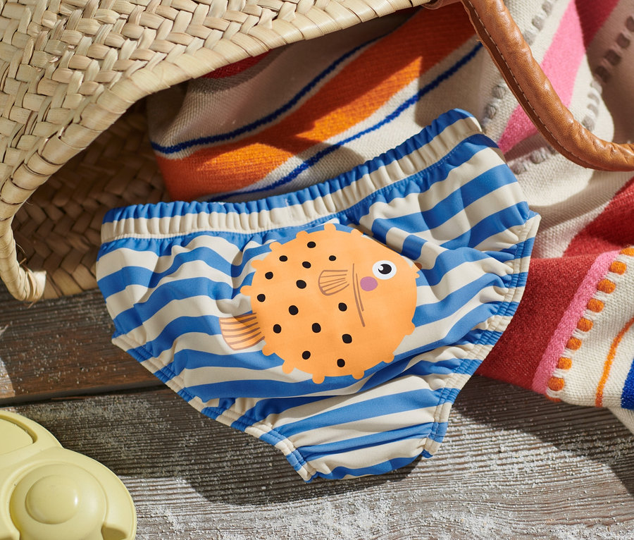 Eine Baby-Schwimmwindel mit blauen Streifen und einem Fischmotiv liegt am Strand neben einem Spielzeug.