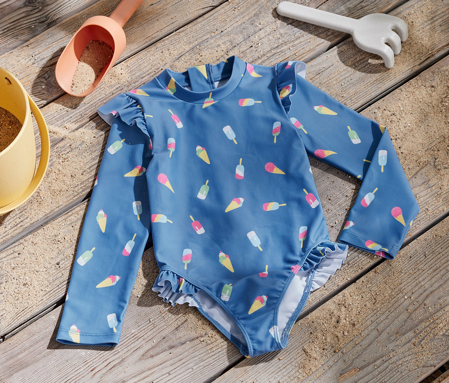 Blauer Kinder-Badeanzug mit Eis-Print und UV-Schutzfaktor 80 liegt neben einem Sandspielzeug auf einem Holzbrett.