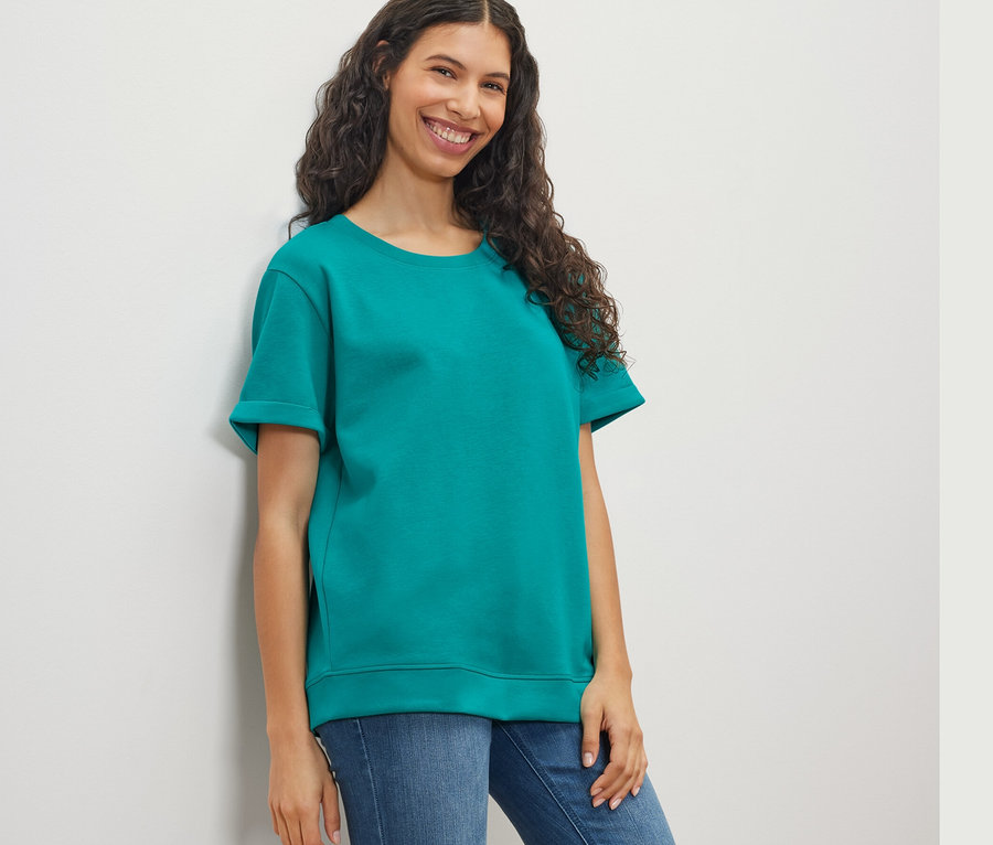 Frau, an eine Wand gelehnt, trägt ein Shirt aus Modalmix in Aqua und Jeans.