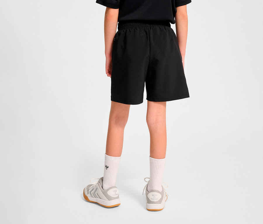 Rückansicht eines Kindes, das schwarze HUMMEL HMLJR Pulse Workout Shorts und Turnschuhe trägt.